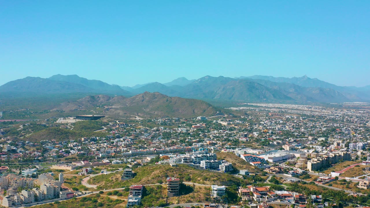 Wide aerial view of San Jose del Cabo with dry hills in the foreground, dense buildings across the middle, blue mountains on the horizon, and no visible people.