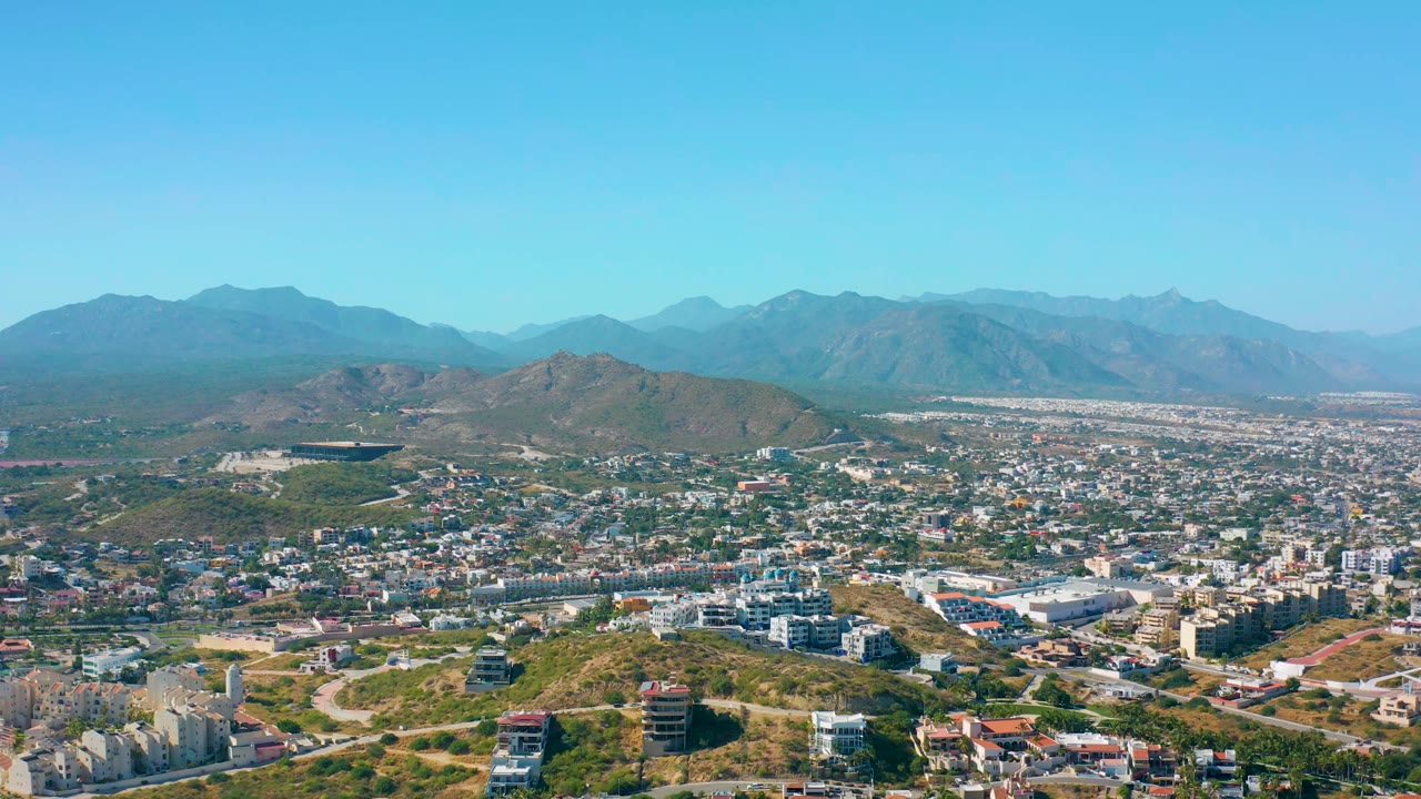San Jose del Cabo remains framed from above with hillside roads and construction in front, a spread of urban buildings in the middle distance, mountains behind, and no visible people.