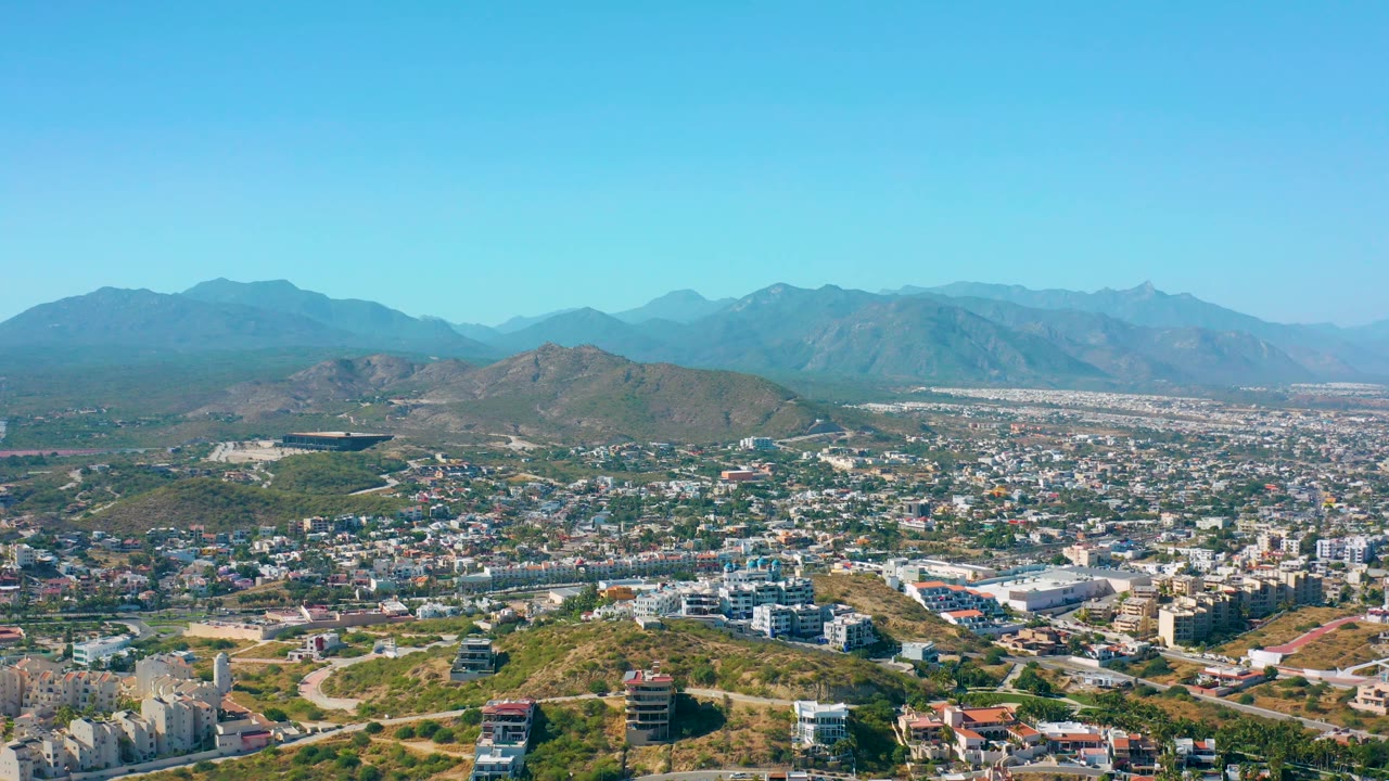 The aerial frame shows San Jose del Cabo stretching across the valley, with low scrub-covered hills in front, compact urban blocks in the center, distant mountains, and no visible people.