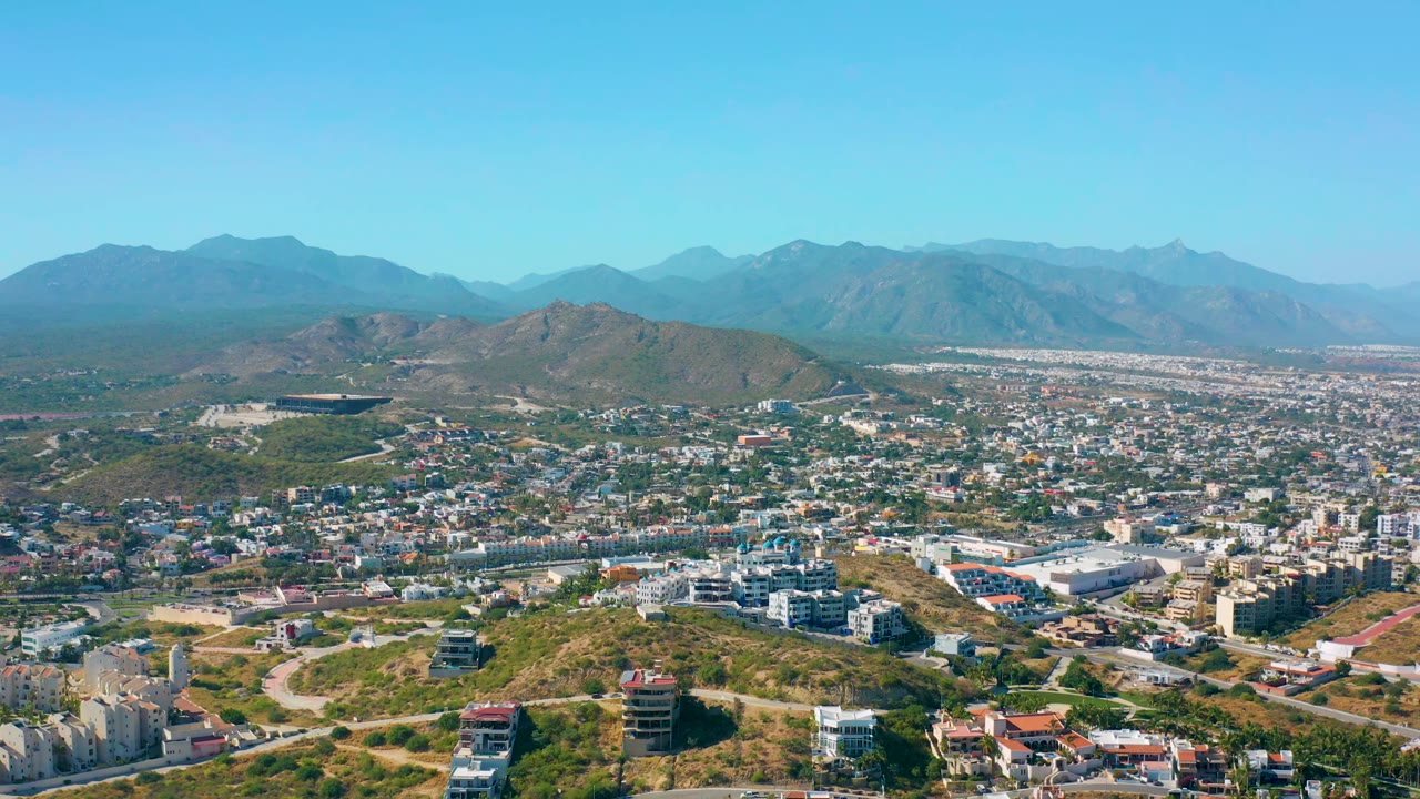 San Jose del Cabo aerial view with dense low-rise buildings in the midground, a rounded hill near center, hazy mountains beyond, and no visible people.