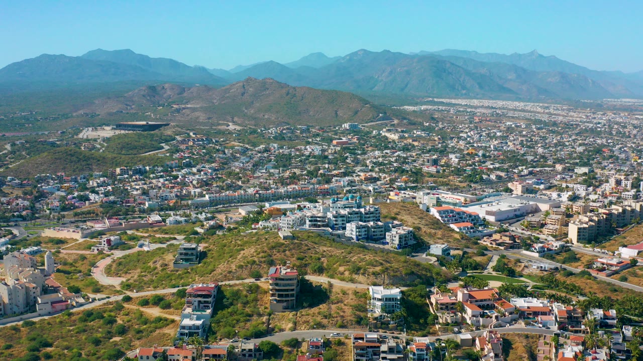 Wide aerial view over San Jose del Cabo with hillside homes, roads, dense urban blocks, distant mountains, clear blue sky, and no visible people.