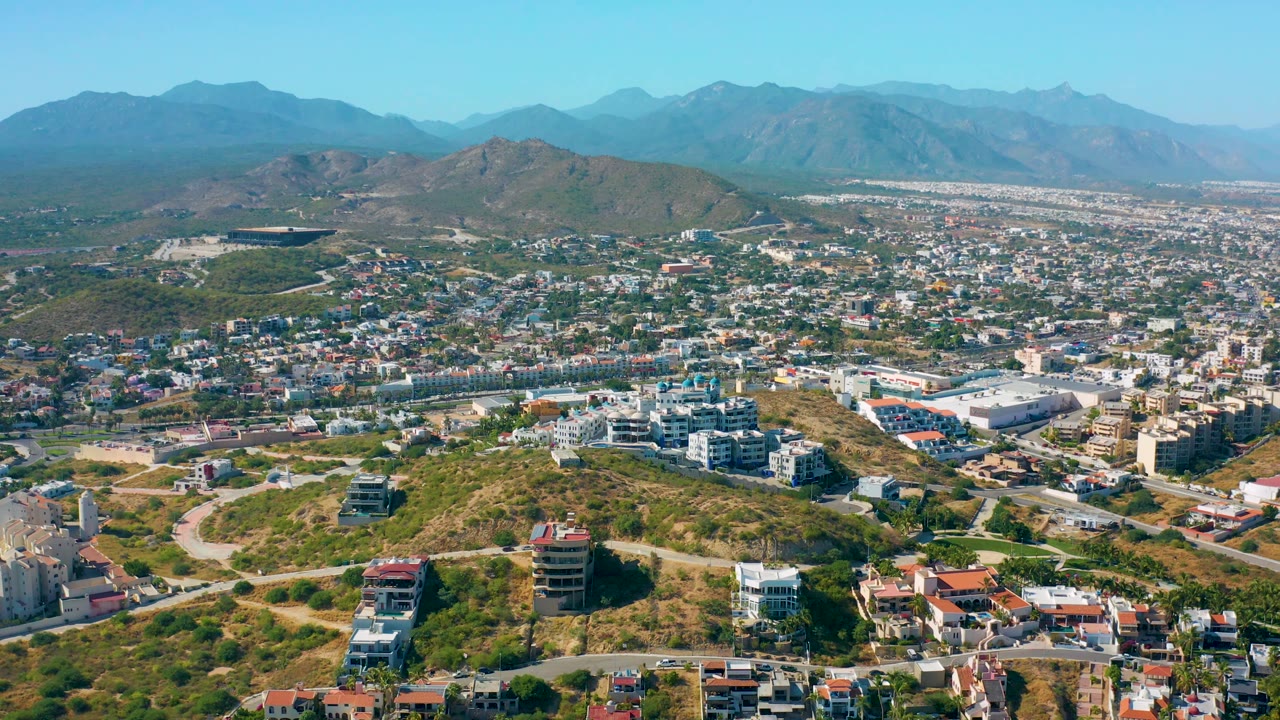 San Jose del Cabo appears from above with a dry hill in the foreground, low-rise city buildings across the center, pale mountain layers in the distance, and no visible people.