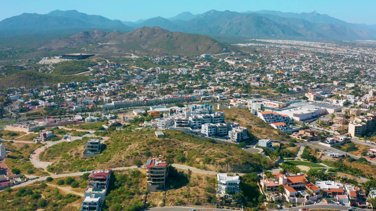 San Jose del Cabo aerial frame with hilltop residences in the lower foreground, a white commercial or civic building cluster in the city below, mountains behind, and no visible people.