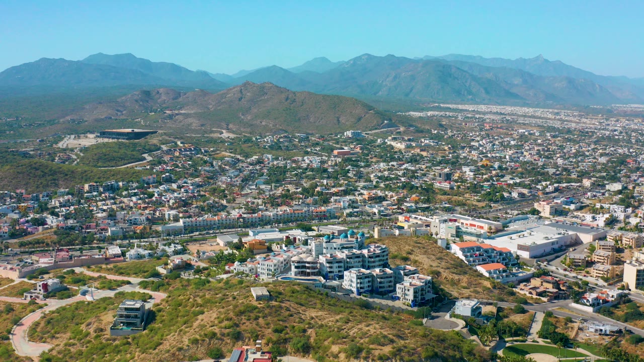 Wide aerial view over San Jose Del Cabo with dry hillside foreground, low-rise city blocks, distant mountain ridges, clear blue sky, and no visible people.