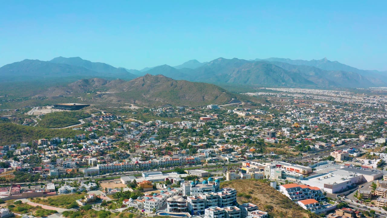 San Jose del Cabo aerial view one second in, showing clustered buildings, road corridors, a rounded hill line, hazy mountains, clear sky, and no visible people.