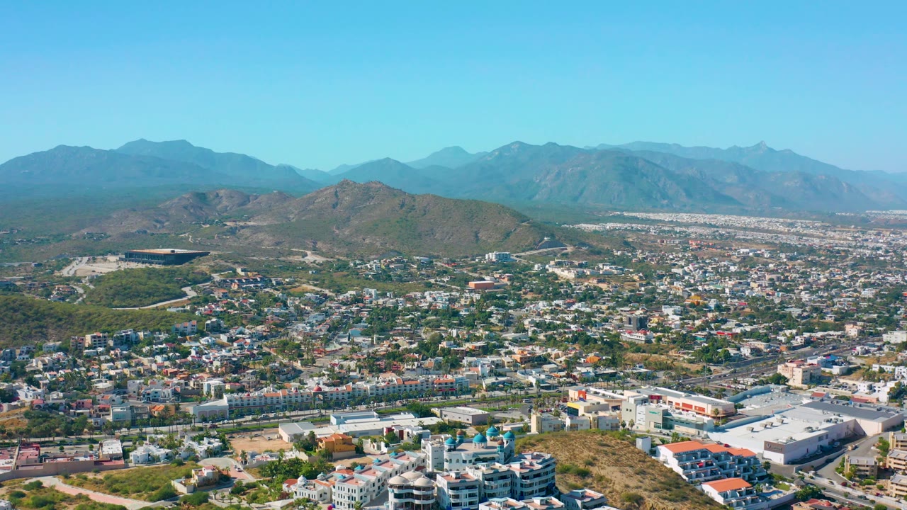 Mid-clip aerial frame over San Jose del Cabo with dense low-rise development in the foreground, a long road network, arid hills, layered mountain ranges, and no visible people.