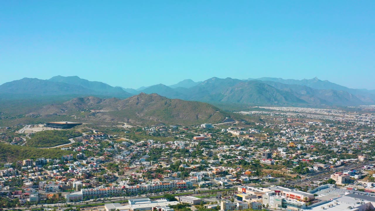 Opening aerial frame of San Jose del Cabo, Mexico, showing low-rise city blocks, green patches, brown hills, a distant mountain range, bright blue sky, and no visible people.