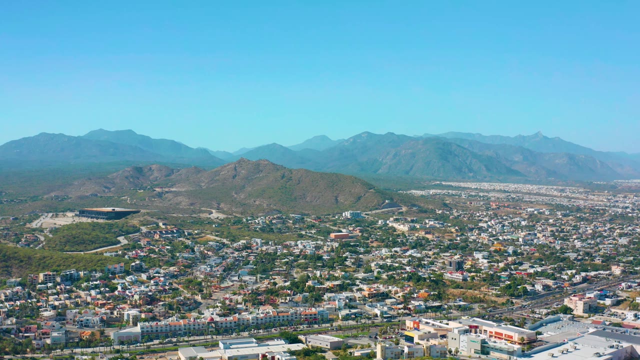 Early frame of the San Jose del Cabo aerial view, with the city grid crossing the lower half, a rounded hill near center, hazy mountains beyond, clear sky, and no visible people.