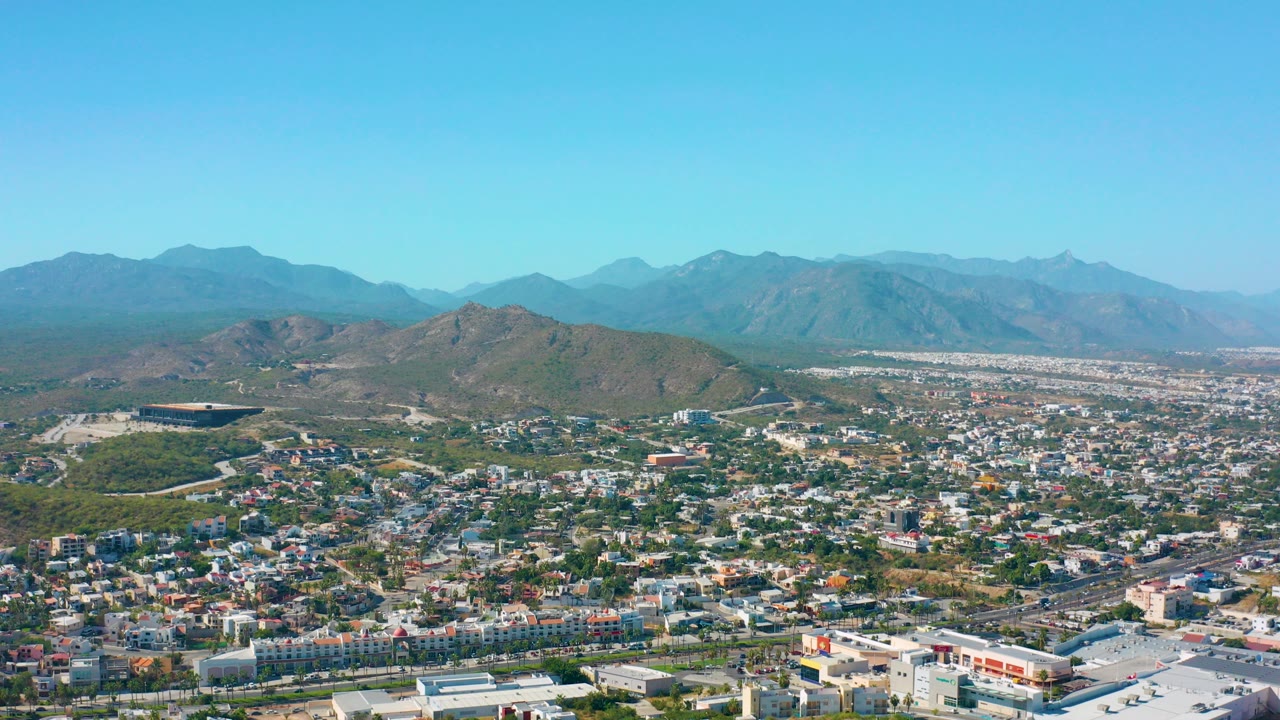 Opening aerial frame over San Jose Del Cabo, Mexico, with low urban buildings in the foreground, green hills beyond, distant mountains on the horizon, and no visible people.