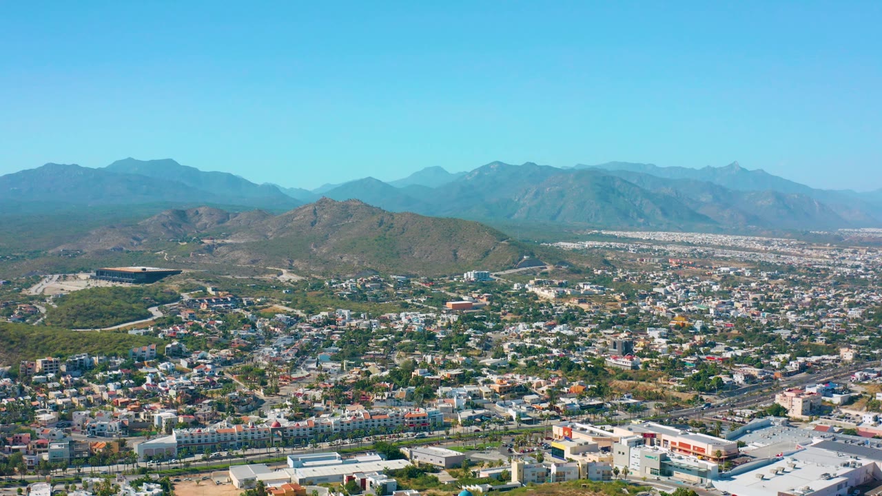 Aerial frame from San Jose Del Cabo, Mexico, with residential and commercial rooftops across the lower frame, rounded hills in the center, distant mountains, and no visible people.