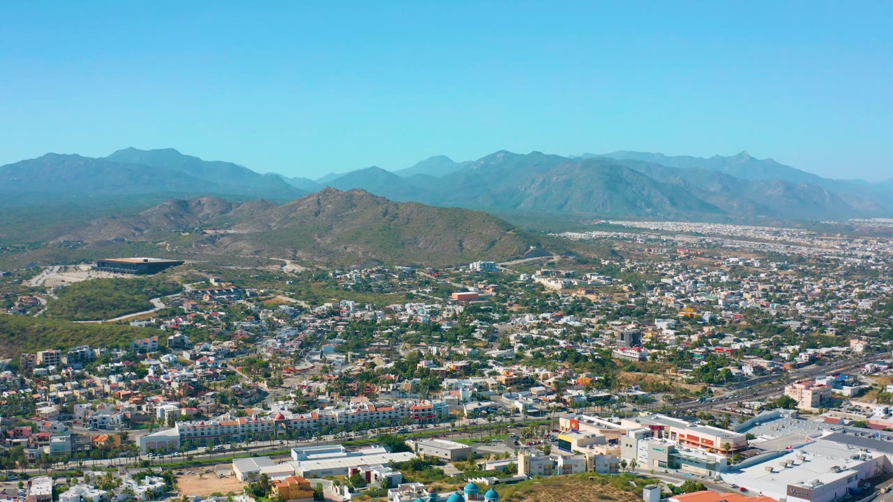 Wide aerial landscape of urban blocks, hillside roads, mountains, and blue sky.