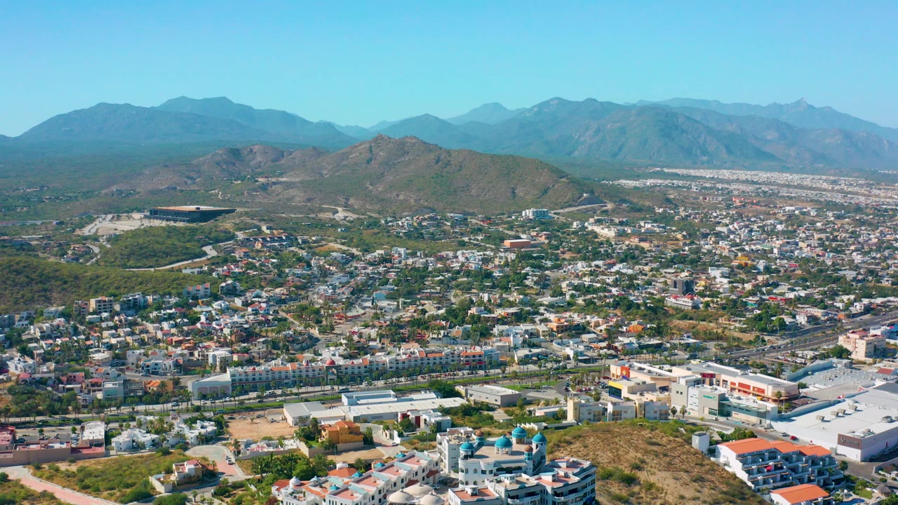 Opening aerial frame over San Jose del Cabo showing low-rise buildings, roads, green hills, distant mountains, and clear blue sky with no visible people.