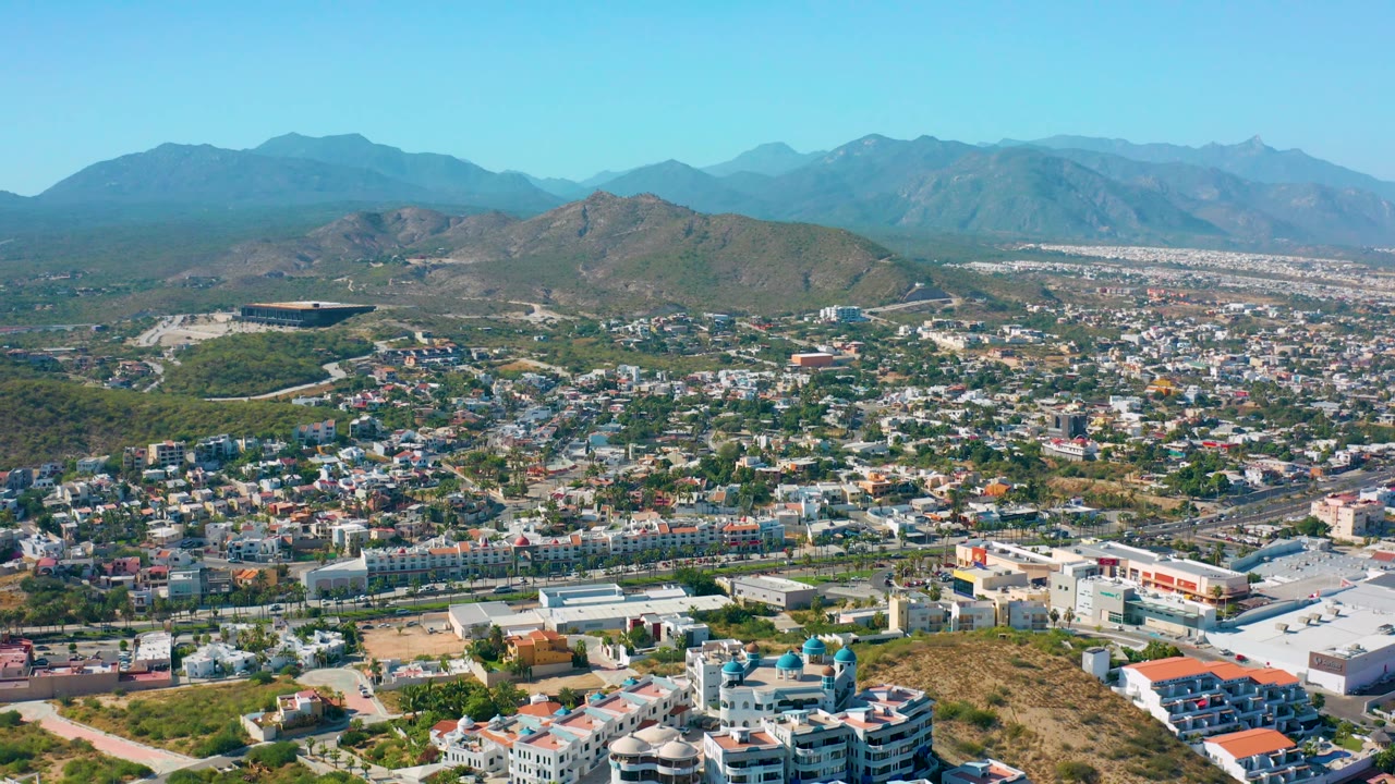 Mid-runtime aerial frame over San Jose del Cabo showing more foreground buildings, a curving road network, green hillside patches, and mountains under clear daylight, no visible people.