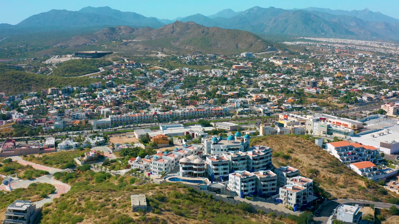 Opening aerial frame over San Jose Del Cabo hillside residences, with dry scrub in the foreground, a dense town beyond, hazy mountains on the horizon, and no visible people.