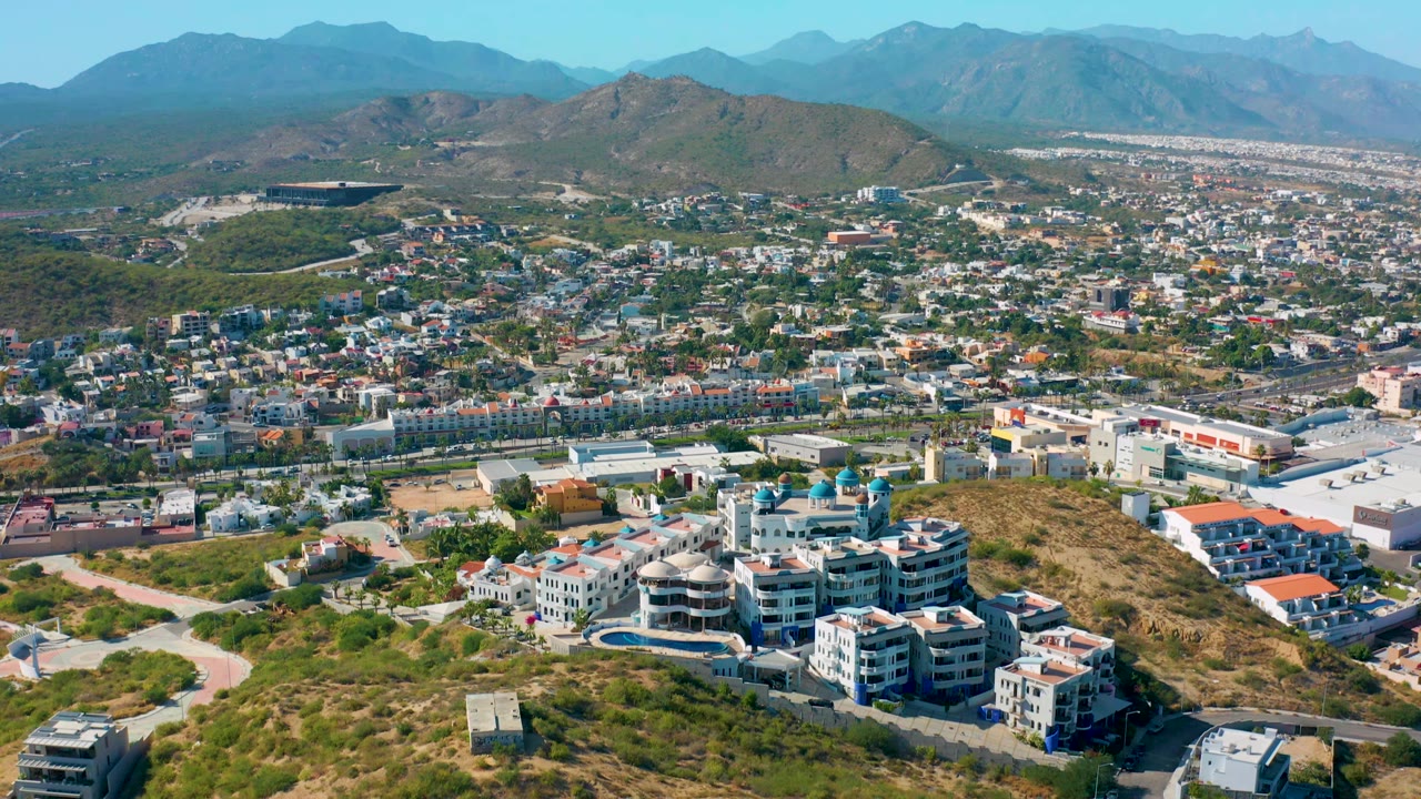 San Jose Del Cabo aerial view with multi-story residential buildings in the lower foreground, dry hillside lots, urban blocks in the distance, mountains, and no visible people.