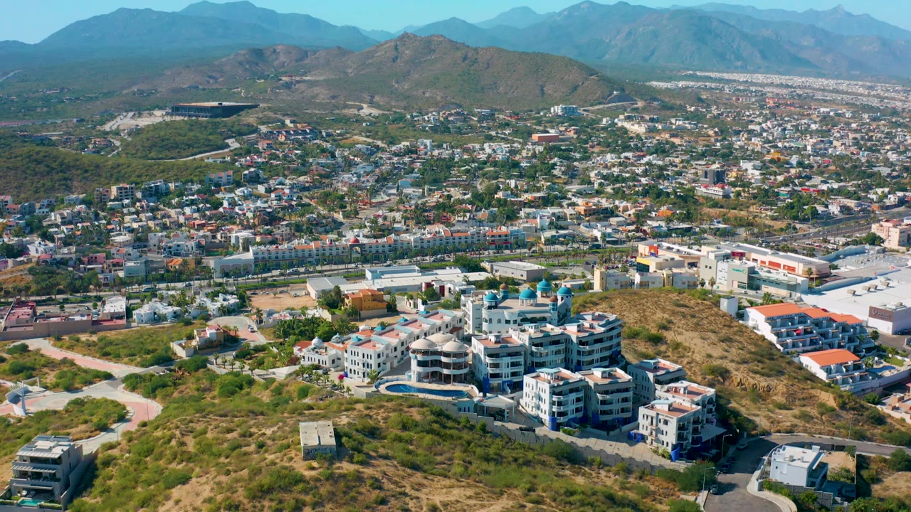 Aerial frame in San Jose Del Cabo showing hillside homes with visible pool areas, curving roads, commercial-size buildings farther back, distant mountains, and no visible people.