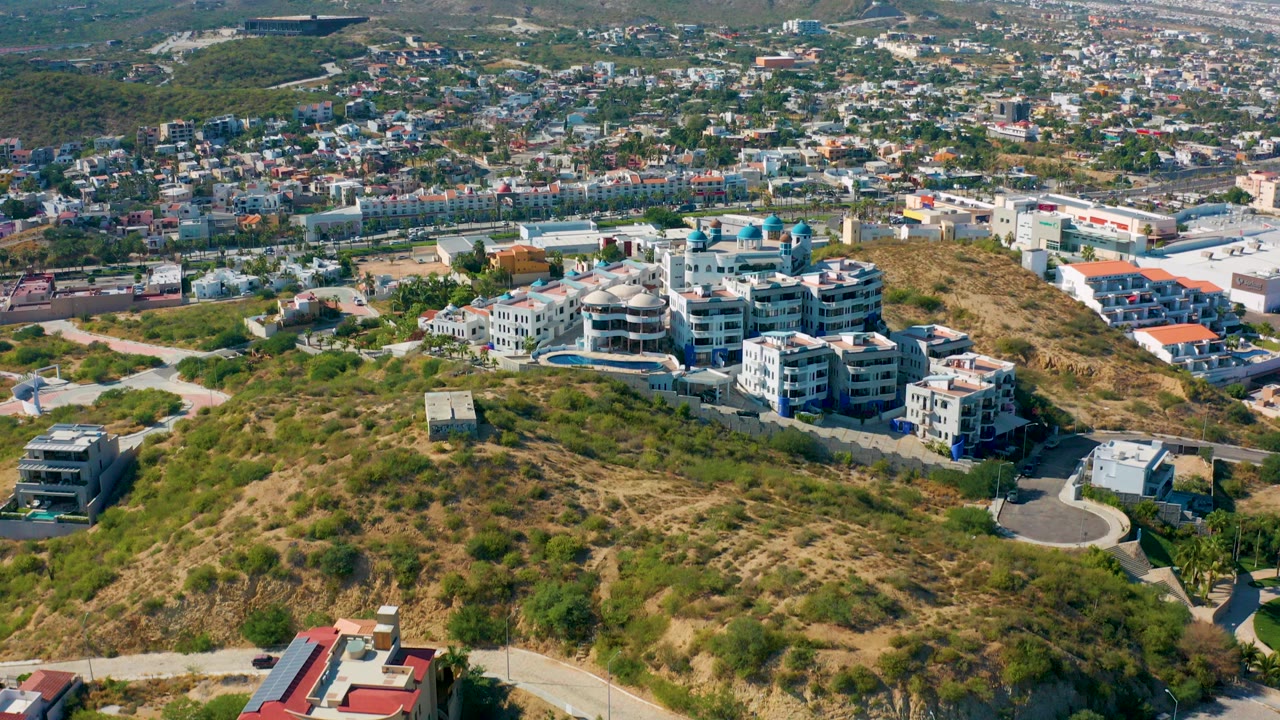 Aerial view of a dry hillside residential area in San Jose del Cabo, Mexico, with curving roads, white apartment buildings, scattered homes, distant hills, and no visible people.