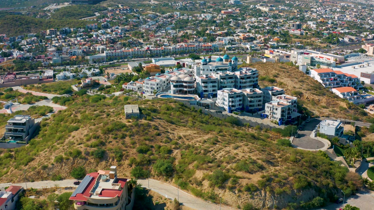 Wide aerial frame of San Jose del Cabo hillside housing, with a scrub-covered slope in front, modern light-colored buildings at center, a road network, background neighborhoods, and no visible people.