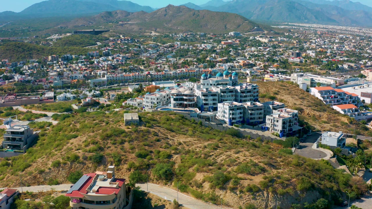 Aerial view over a San Jose del Cabo hillside with modern homes, dry vegetation, city development, distant mountains, blue sky, and no visible people.