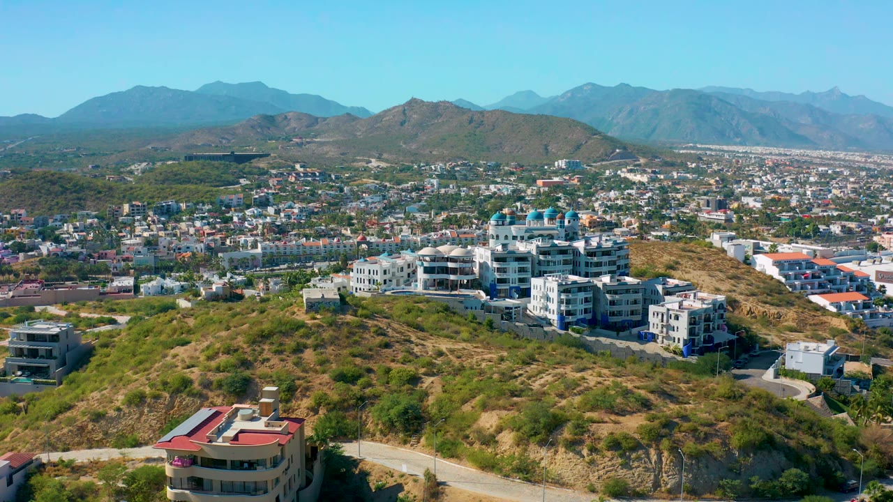 Aerial view in San Jose del Cabo with dry green hillside in front, clustered white buildings in the midground, distant city blocks, mountains, and no visible people.