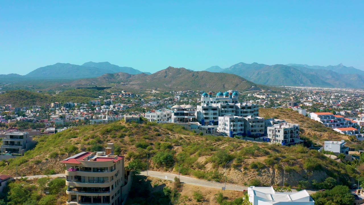 San Jose Del Cabo hillside view with a foreground road, multi-story residential buildings, distant city blocks, mountains, blue sky, and no visible people.