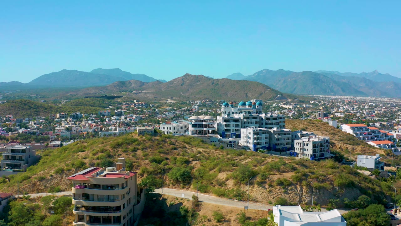 San Jose Del Cabo aerial frame with white hilltop buildings centered, a tan road across the slope, spread-out town blocks, mountains, and no visible people.
