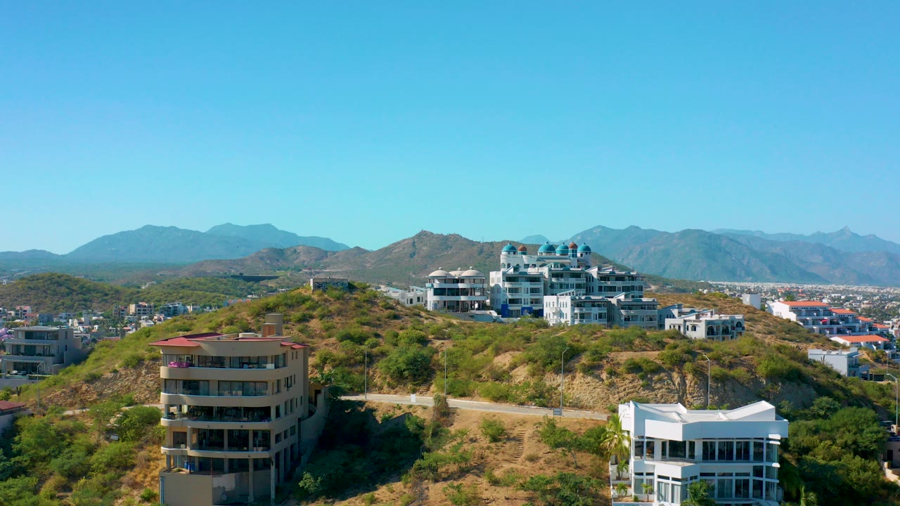 Opening frame of San Jose Del Cabo hillside residential buildings, dry scrub slopes, distant mountains, and no visible people under a clear blue sky.