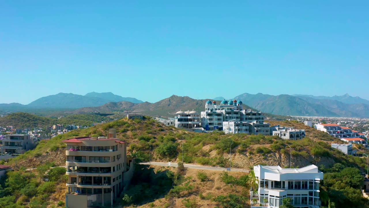 Residential buildings on a dry green hillside with mountains beyond