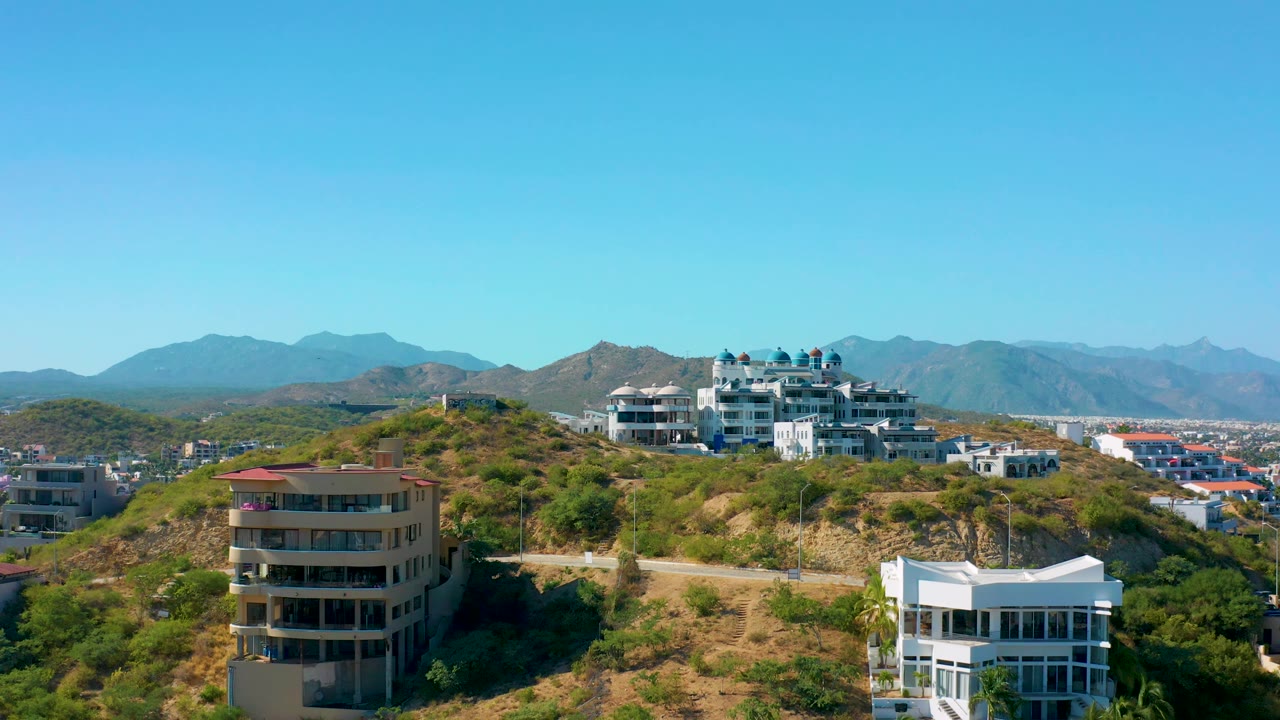 The aerial view holds on modern white residences, a scrub-covered hill, distant ridgelines, and a clear sky in San Jose Del Cabo, with no visible people.