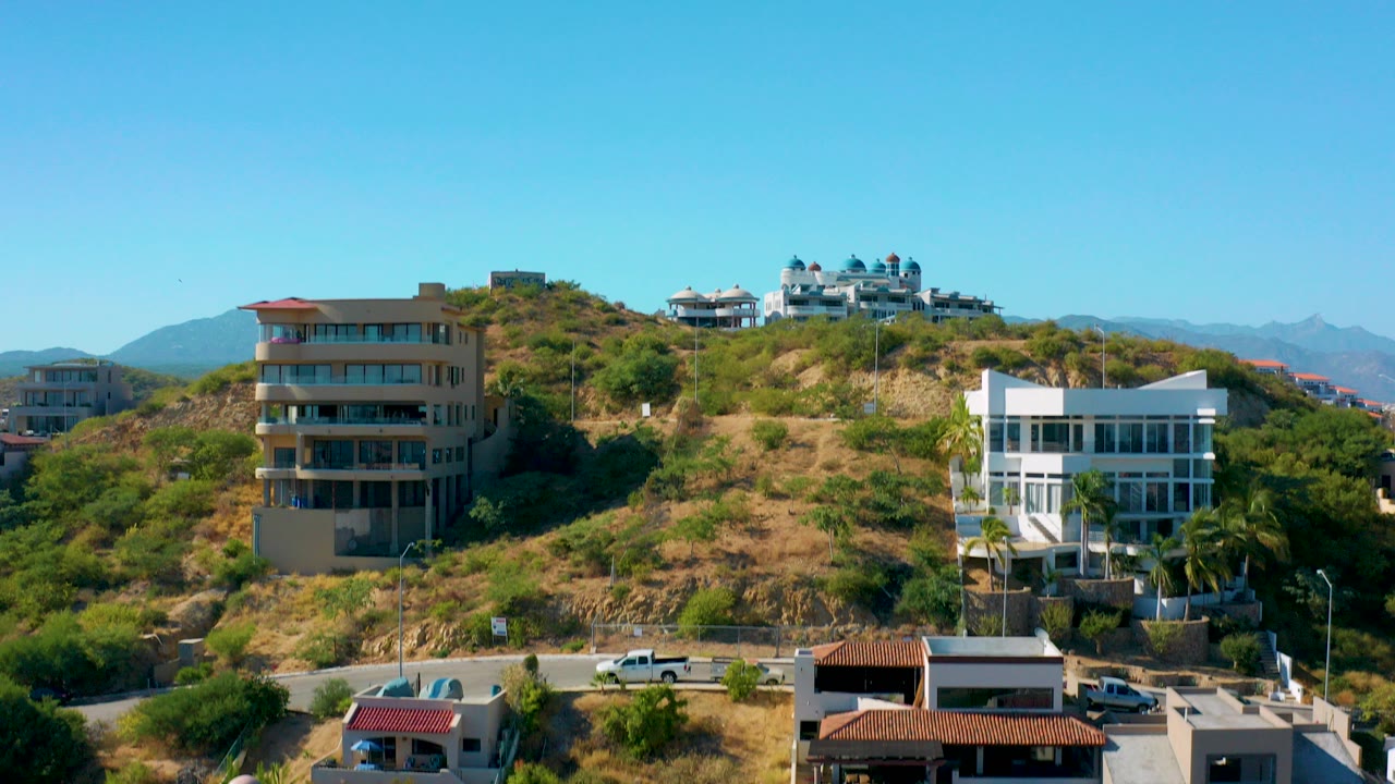 San Jose Del Cabo hillside with modern residential buildings, dry grass, shrubs, distant mountains, parked vehicles, and no visible people at the start of the aerial view.