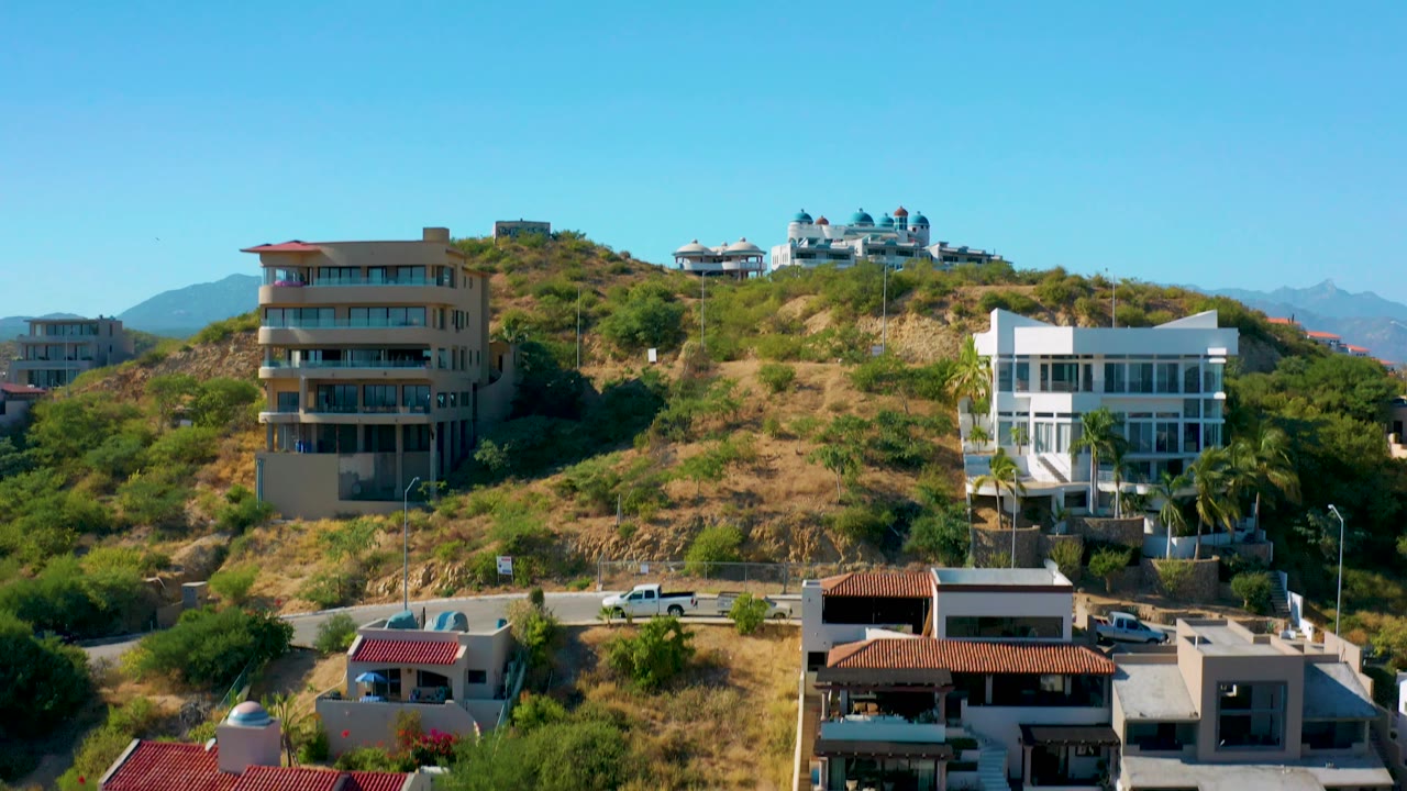 San Jose Del Cabo hillside residences fill the frame, with dry scrub, a white multi-story home, an apartment block, parked vehicles, and no visible people.