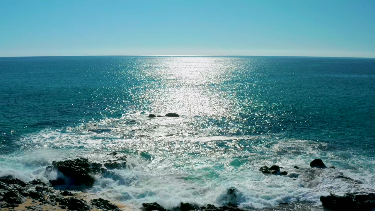 San Jose del Cabo ocean view with foamy water spreading around black rocks, a bright vertical path of reflected sunlight, and no visible people.