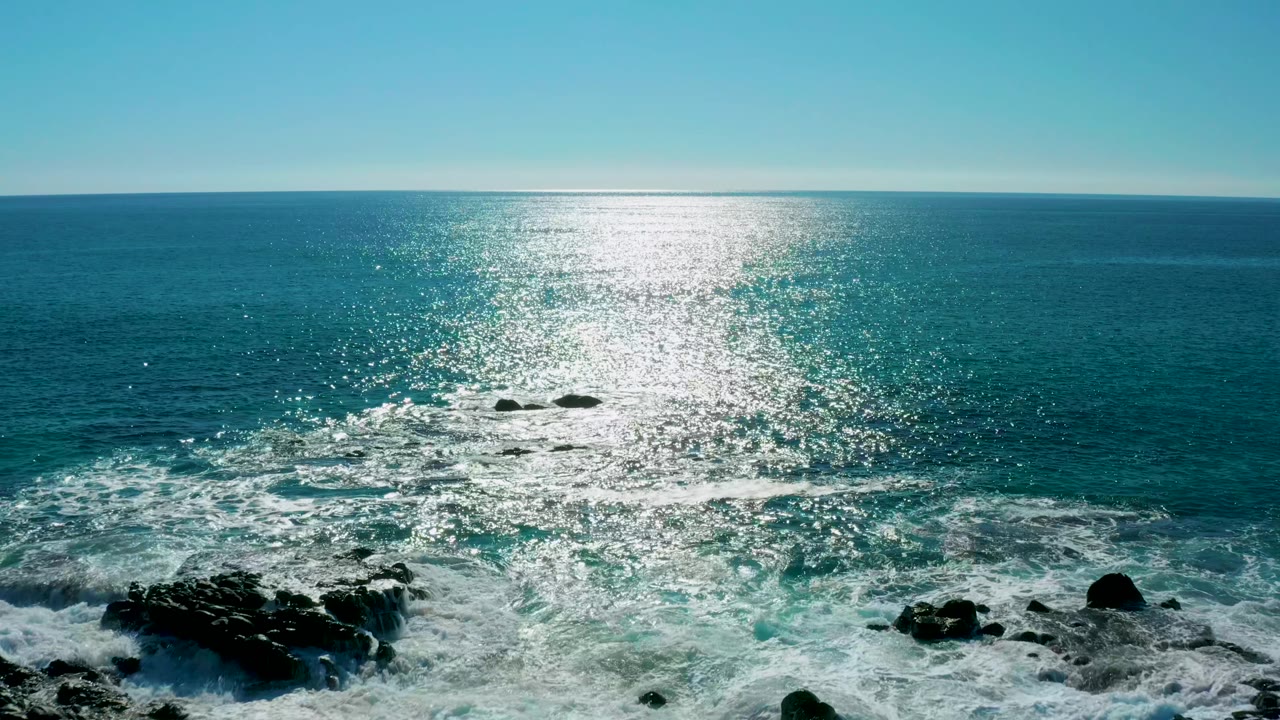 Elevated view of the San Jose del Cabo coastline with churning white surf over dark rocks, sparkling blue water beyond, and no visible people.