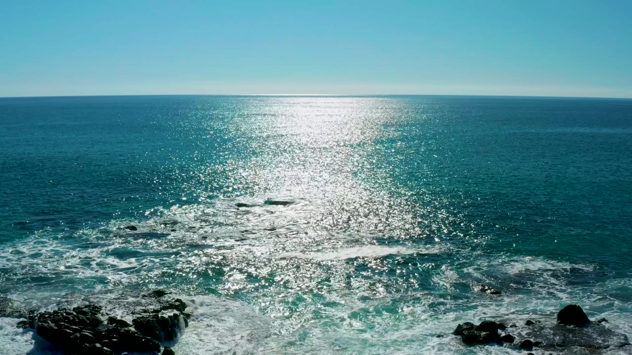 Wide ocean surface with waves breaking near rocks at lower left
