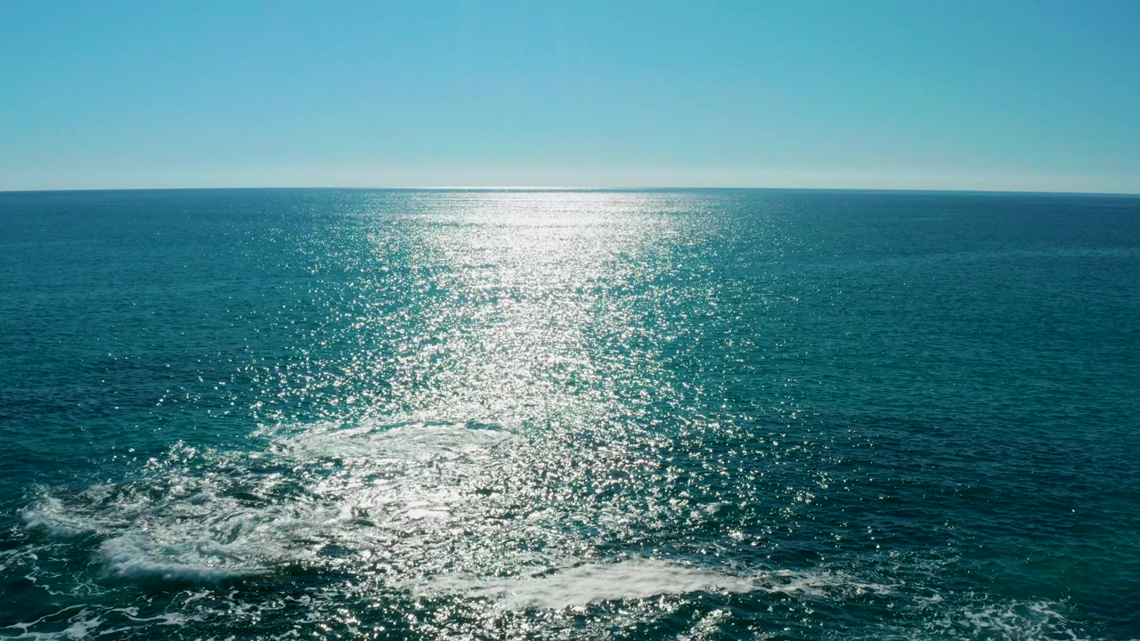 Sunlit blue ocean with white wake foam near rocks