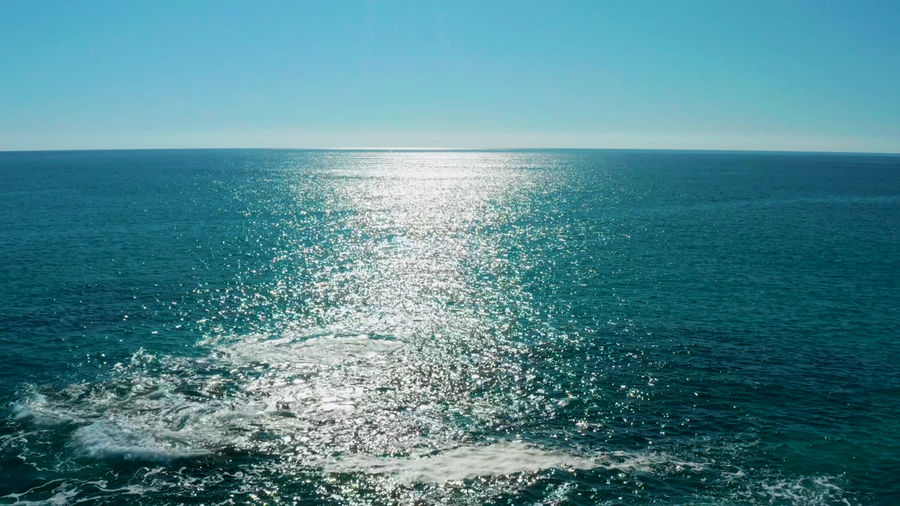 A wide aerial view in San Jose Del Cabo shows the horizon, turquoise-blue water, a bright reflection column, white foam drifting over submerged rocks, and no visible people.