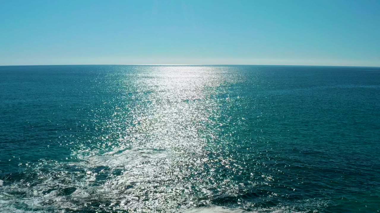 Sunlight glitters across open ocean in San Jose Del Cabo while white wake foam curls near dark rocks in the lower frame, with no visible people or vessels.