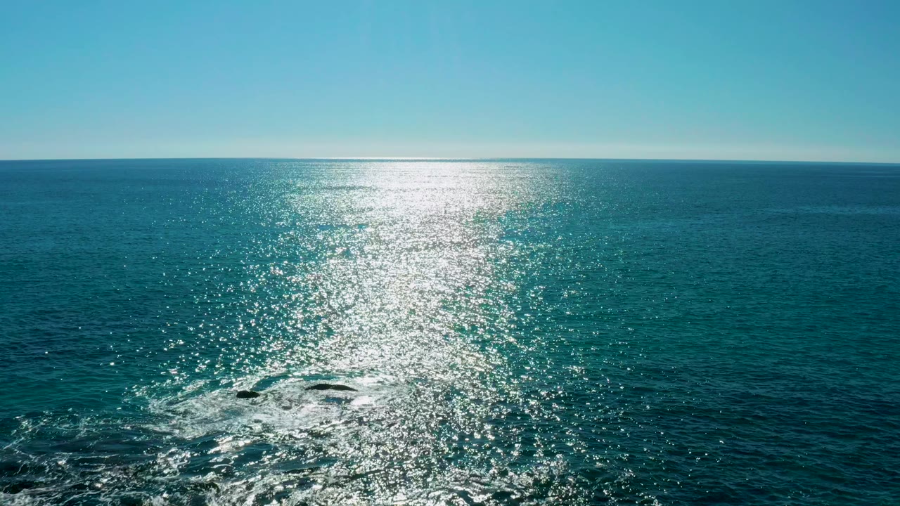 The San Jose Del Cabo ocean fills the frame with a level horizon, a tall band of sun glare, faint wake foam near lower-left rocks, and no visible people.