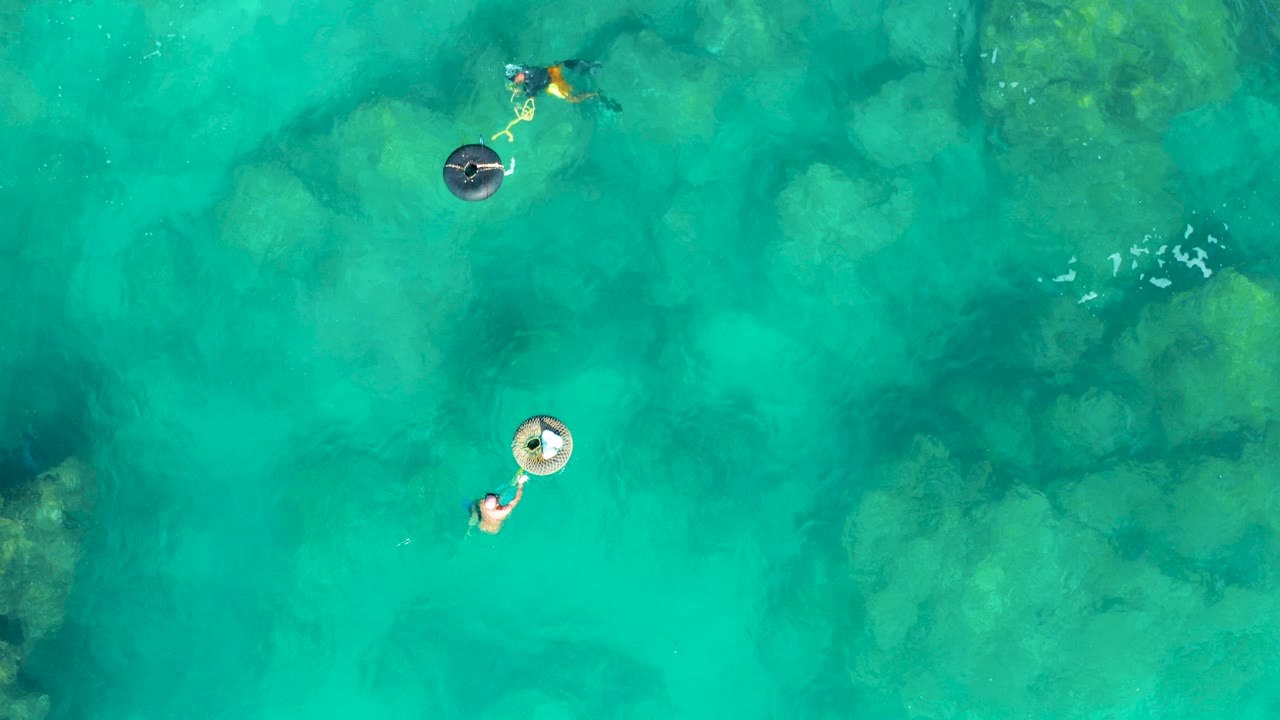 Overhead frame from San Jose Del Cabo showing two people separated in clear turquoise water, one near a pale inflatable ring and one near a dark ring over submerged rocks.
