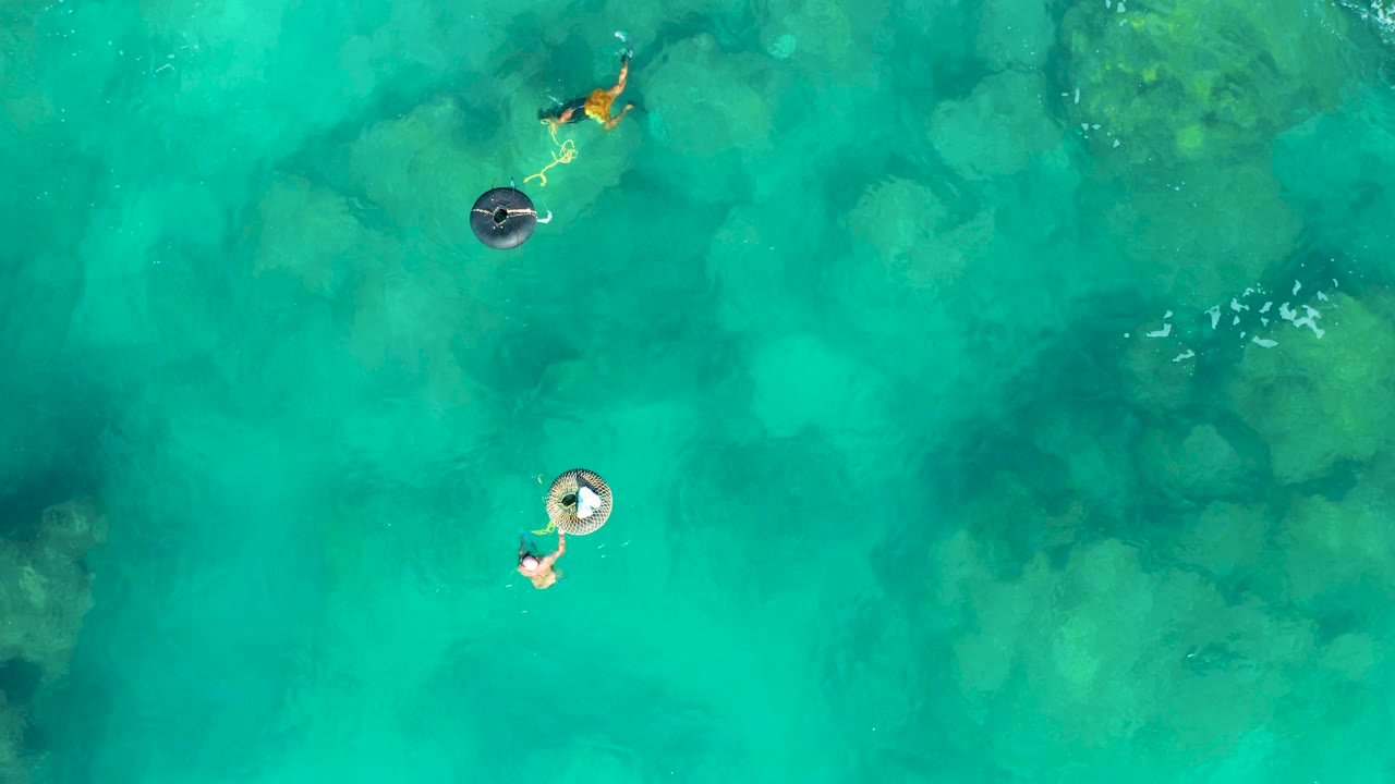 At 0:02 in San Jose Del Cabo, the overhead view keeps both people and their inflatable rings near the left side while broad green-blue water and submerged rocks fill the rest of the frame.