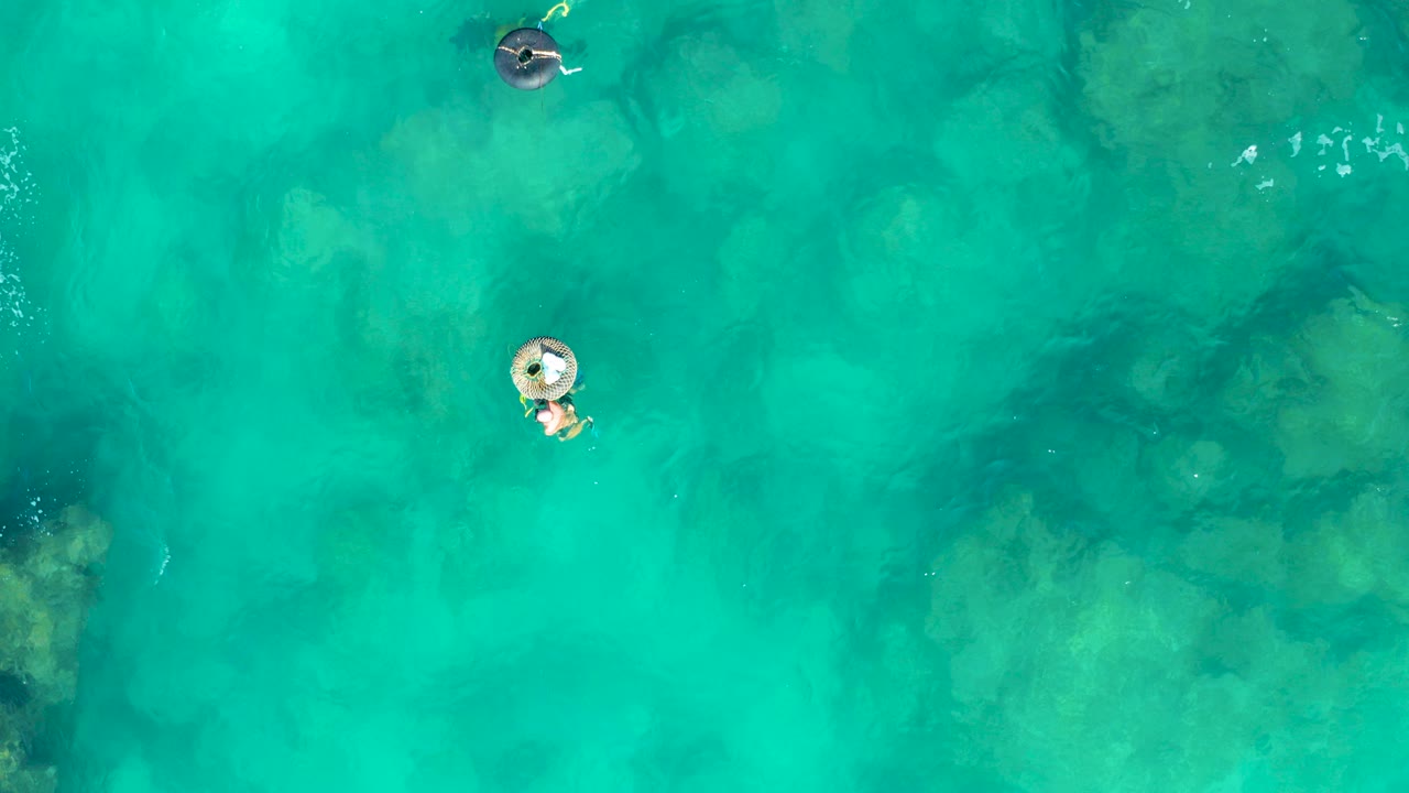 Overhead view in San Jose Del Cabo of two people on inflatable rings separated across clear turquoise water, with darker seabed patches visible below.