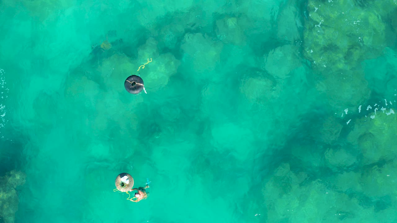 Overhead frame from San Jose Del Cabo shows two people floating separately on inflatable rings over clear turquoise water with underwater rocks visible below.