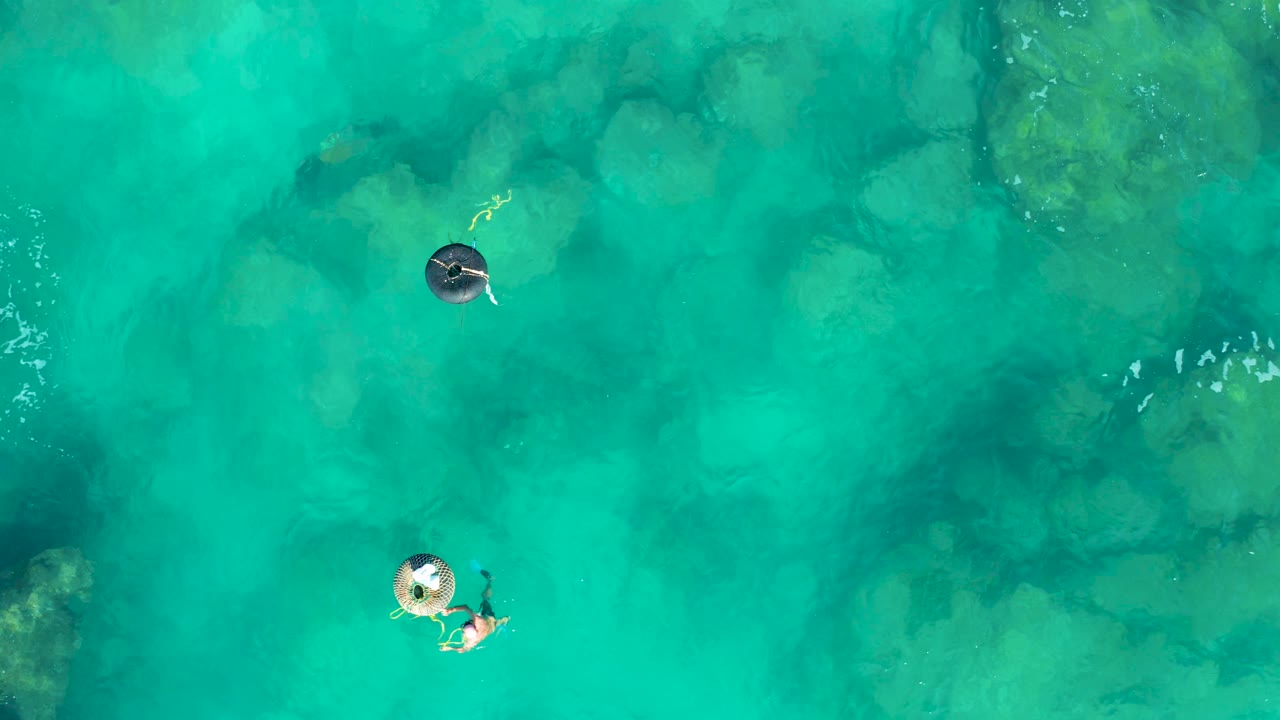 In this overhead San Jose Del Cabo frame, two people on rings float across open turquoise water while submerged rocks form darker green patches below.