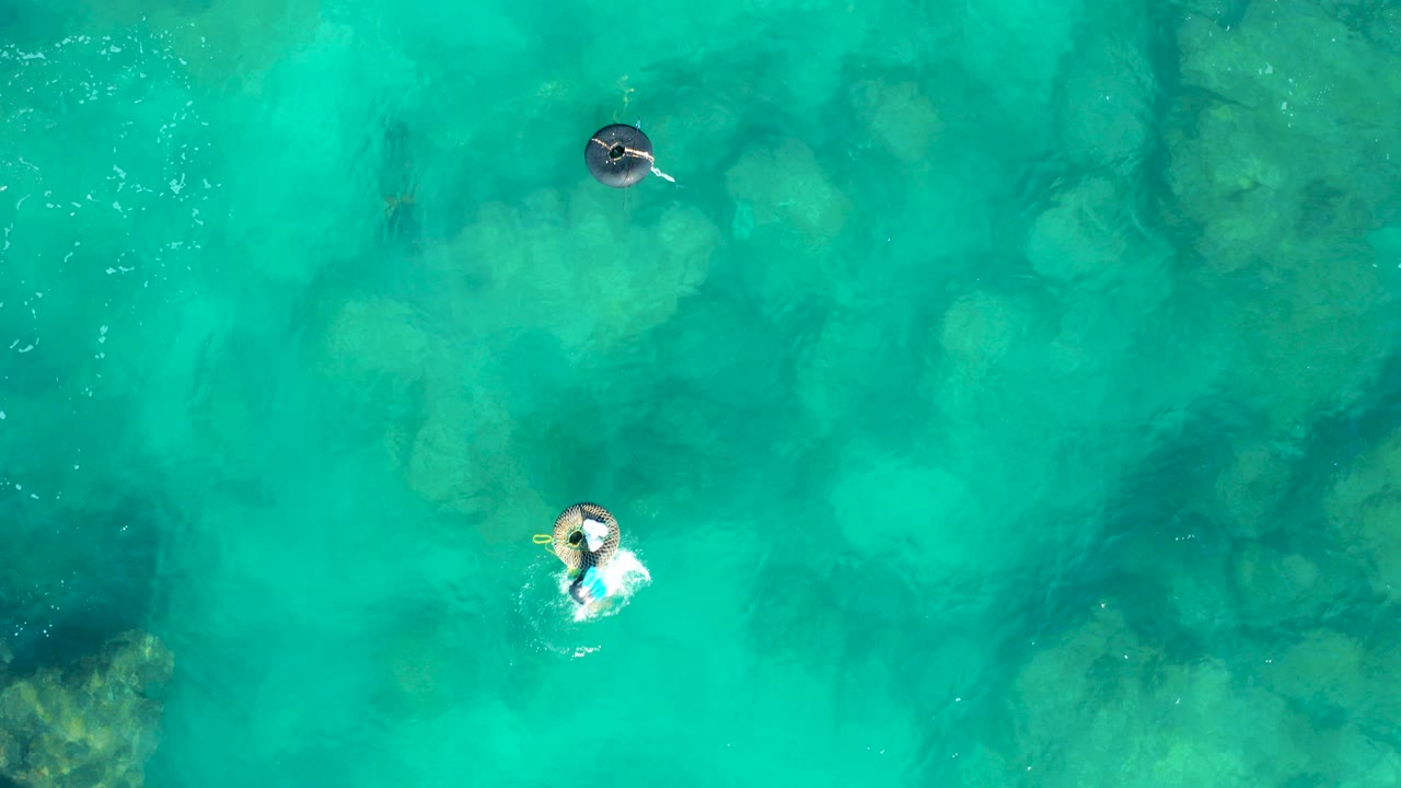 Overhead view in San Jose Del Cabo of two people floating apart on inflatable rings above clear turquoise water with submerged rocks and light ripples.