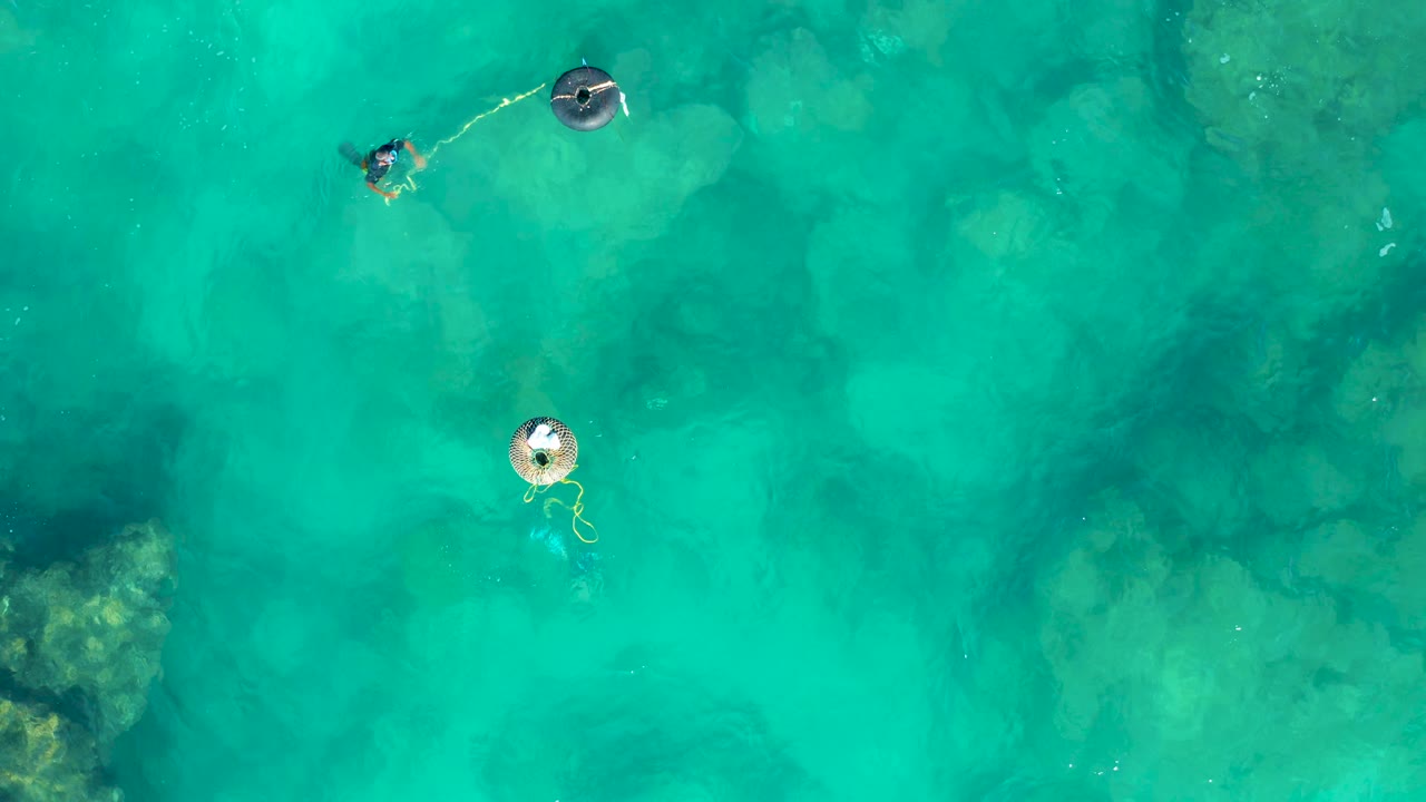 At 0:02, one person trails beside a dark inflatable near the top of frame while another remains near a light ring over clear water in San Jose Del Cabo.