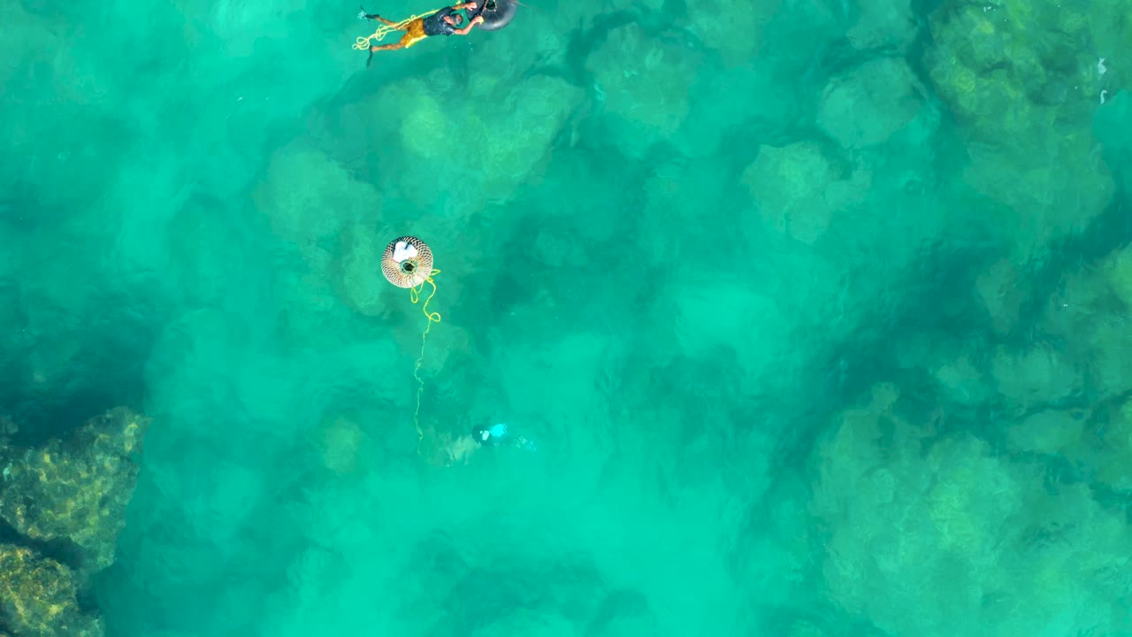 Aerial frame over San Jose del Cabo water places the float near the left center, a small swimmer below it, and another swimmer near the top edge above pale submerged rocks.