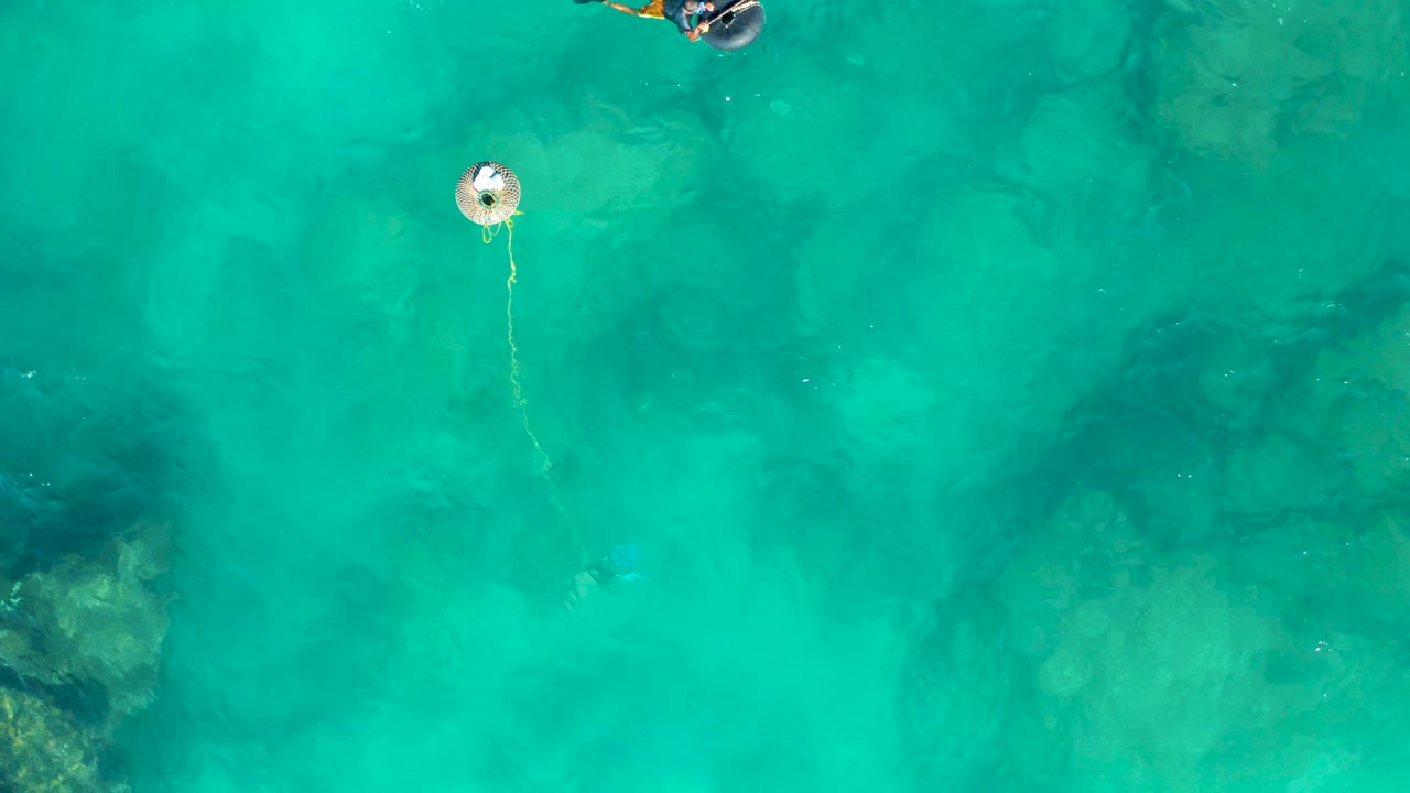 Top-down San Jose del Cabo view with the snorkeler near the upper edge, a round buoy below, and darker submerged rock shapes visible through turquoise water.