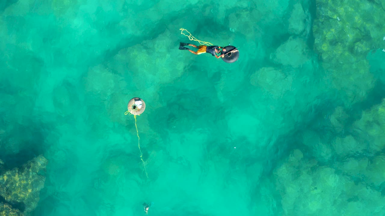 Overhead view in San Jose del Cabo of two snorkelers near the top of frame, a swimmer lower in the water, and a white buoy with rope over turquoise water and rocks.