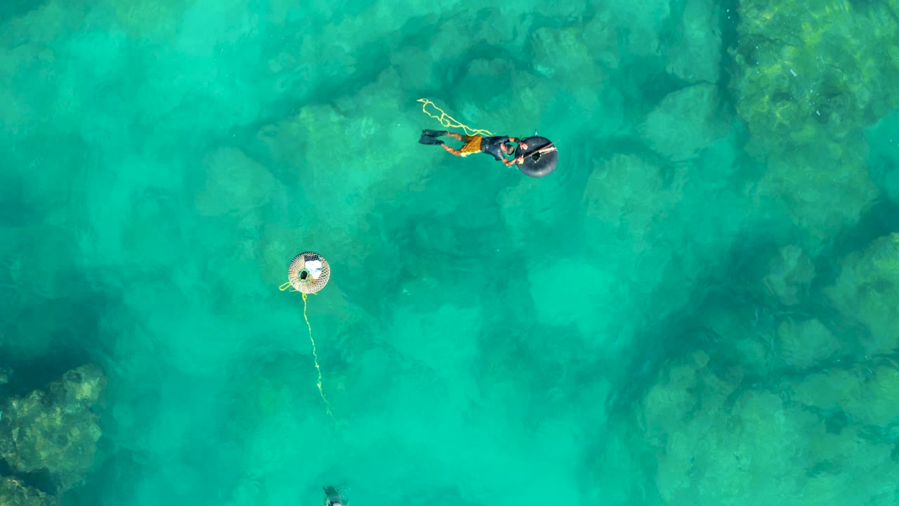 Overhead San Jose del Cabo frame with two snorkelers at the upper right, a white buoy below them, and dark underwater rocks showing through bright turquoise water.