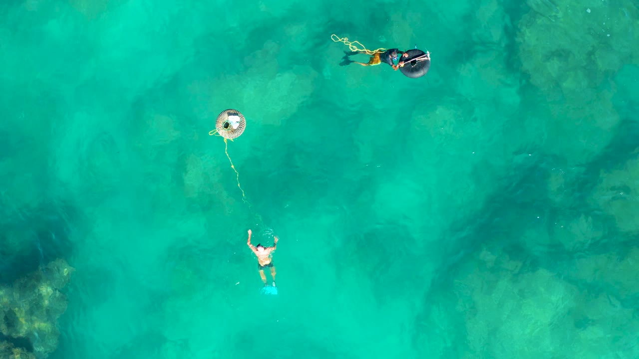 Top-down San Jose del Cabo water view where the lower swimmer raises both arms while the snorkeler remains near the upper edge and the buoy sits between them.
